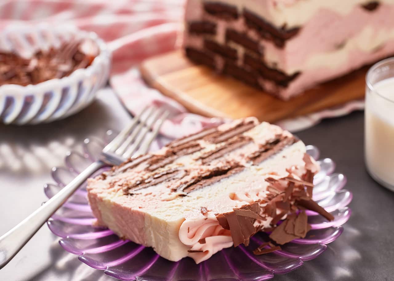 A retro scalloped purple glass plate holds a slice of ice-box xake, with chocolate biscuits sitting in cream and pink-coloured cream layers. A fork leans on the plate. Uncut cake sits on a serving board in the background.  