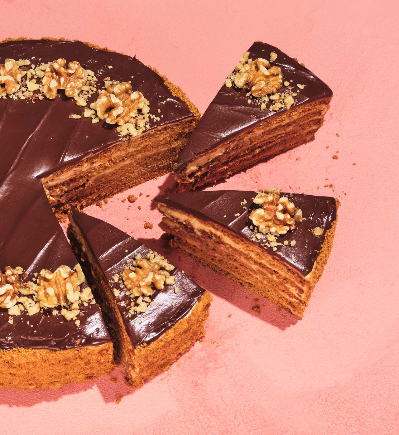 A round cake topped with chocolate icing and walnut halves sits on a peachy-red surface. Three slices have been cut from the cake and sit slightly away from it, showing the cake layers within. 