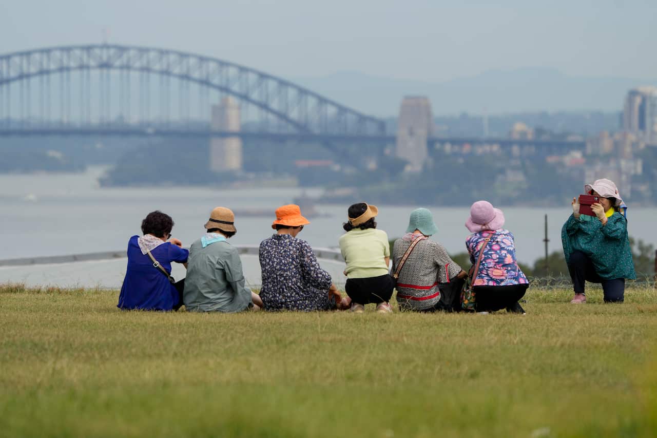 A group of women wearing pastels and hats sit on the grass overlooking the ocean and the Sydney Harbour Bridge. A woman takes a picture on her phone of the group.