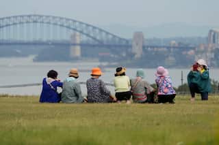A group of women wearing pastels and hats sit on the grass overlooking the ocean and the Sydney Harbour Bridge. A woman takes a picture on her phone of the group.