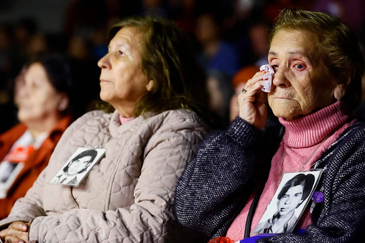 Two women sitting next to one another. One woman is drying her eye with a handkerchief. Both women have black-and-white pictures of people pinned to their front