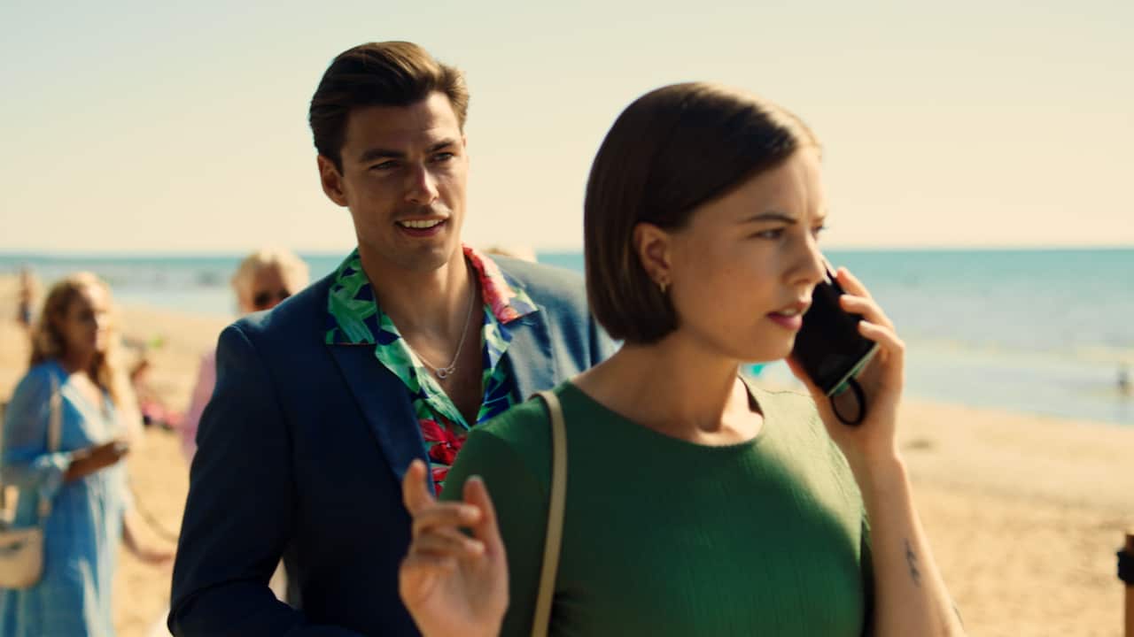 A woman in a green dress stands on a beach, talking into a phone. A man in a bright shirt and blue jacket stands behind her as if about to speak to her. 