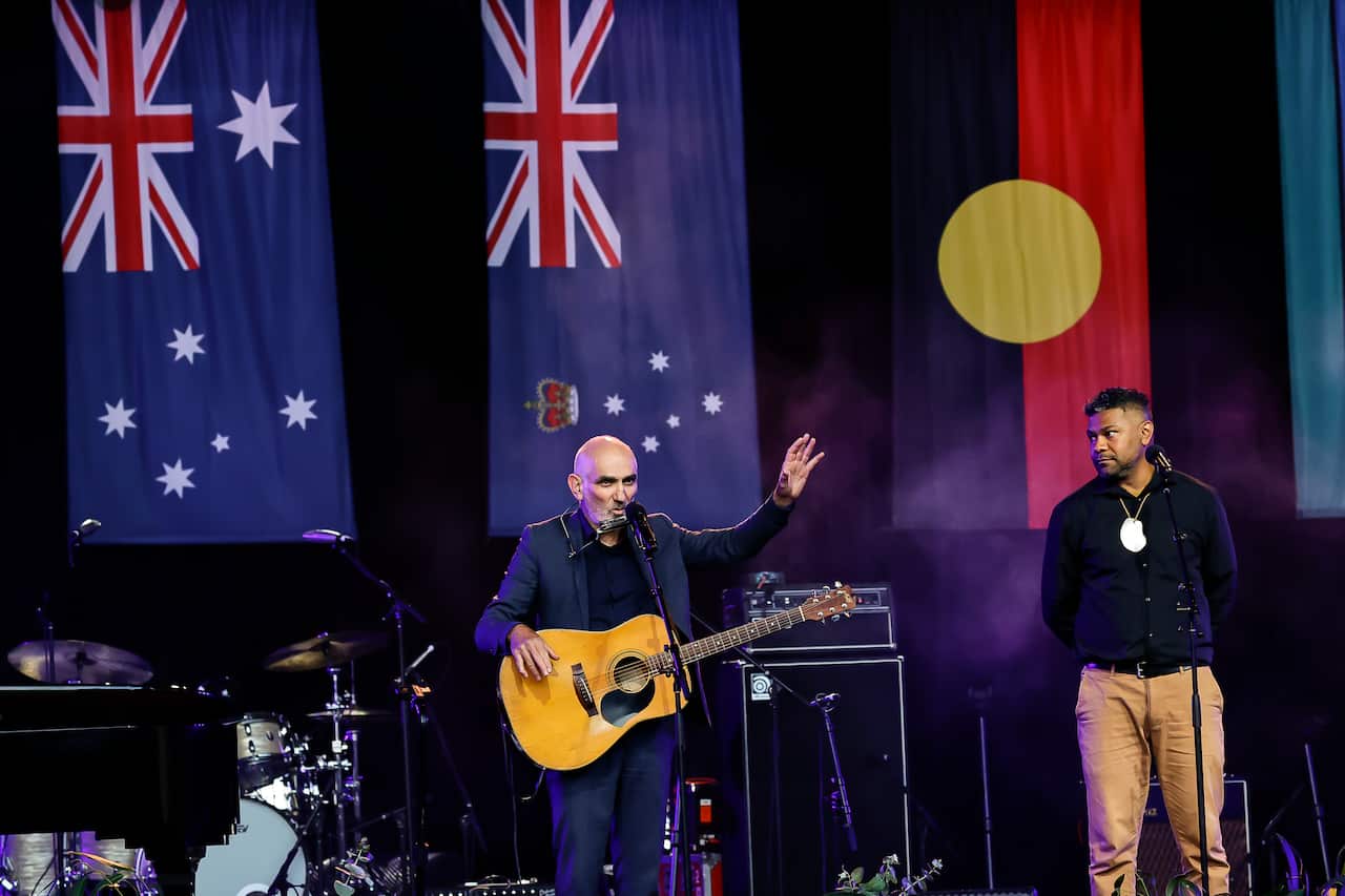 two men stand on stage in front of microphones, one holding a guitar, with Australian and Aboriginal flags draped from the ceiling behind them.
