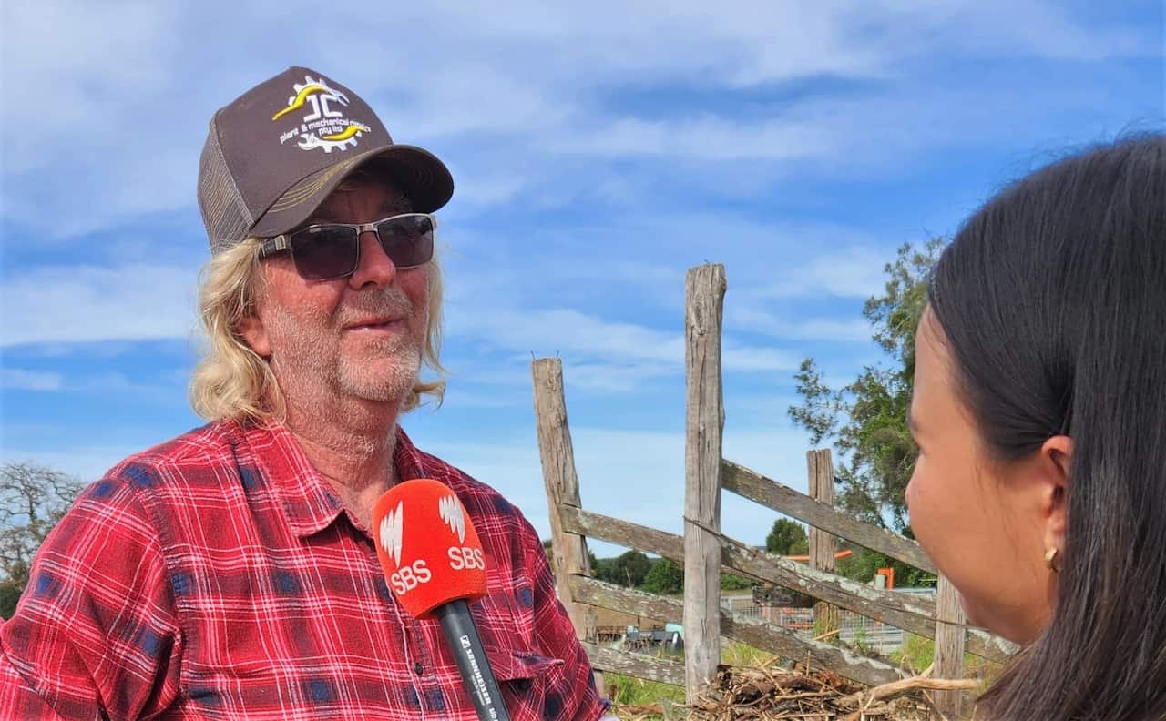 An older man with shoulder-length white hair, wearing a black graphic cap, and red tartan shirt and sunglasses. He is speaking to a dark haired reporter holding up a red SBS News microphone. In the background is a farm scene with blue sky above. 