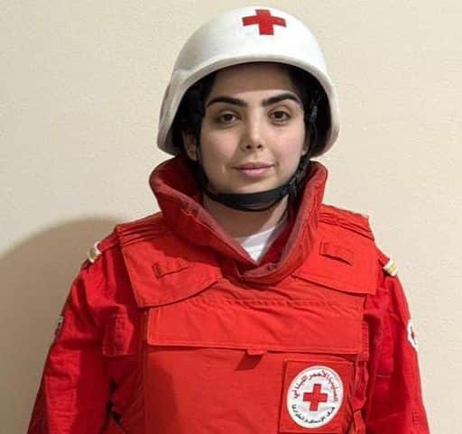 A woman in a Red Cross paramedic uniform, wearing a white helmet, stands in front of a wall.