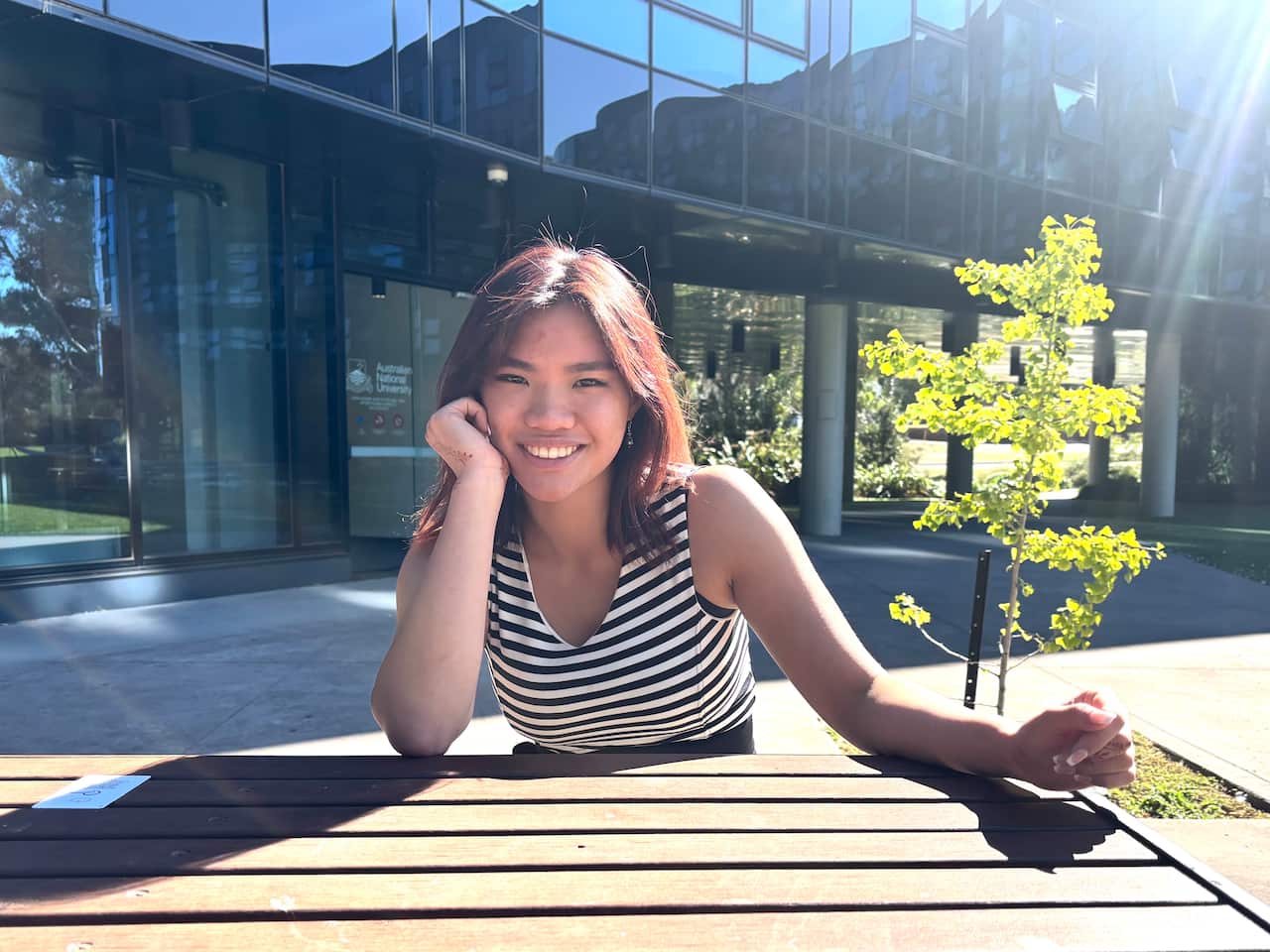 A woman with brown hair and wearing a black and white horizontal striped tank top sits at a wooden table outside and smiles for the camera