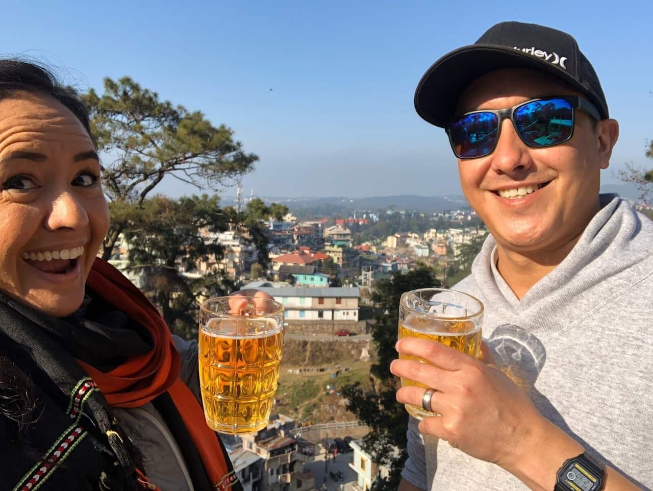 A man and woman holding beers and smiling, a town is in the background. 