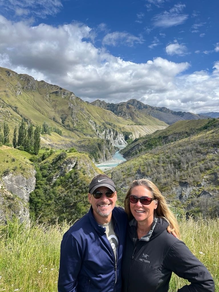A man and woman both wearing sunglasses and raincoats smile in front of a river and mountains. 