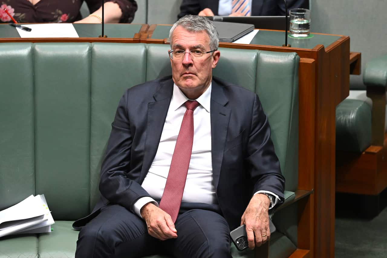 A man wearing a suit and tie sits on a sofa, holding a phone in his left hand.