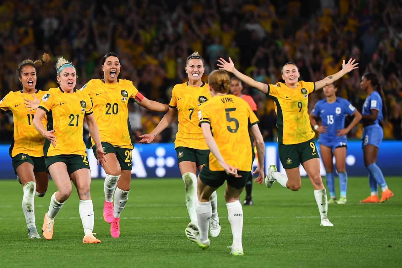 A woman in a yellow football uniform and pink boots, with two women in blue football uniforms on her left and right sides.