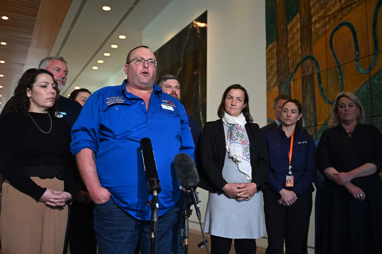 A man in a blue shirt speaking in front of microphones in a hallway. Other men and women surround him.