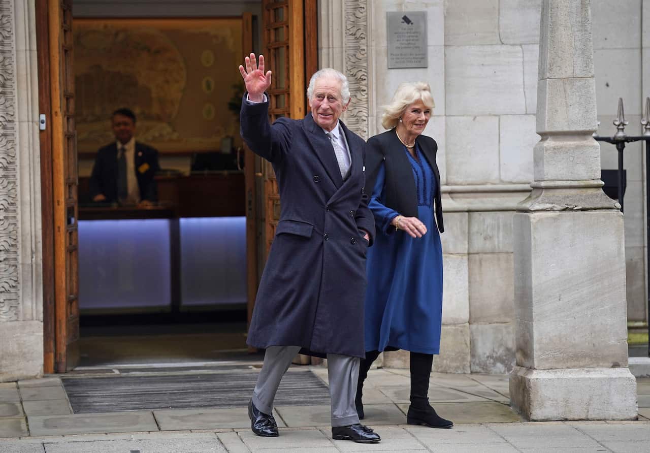 King Charles, wearing a black coat, and with Queen Camilla by his side, waves.