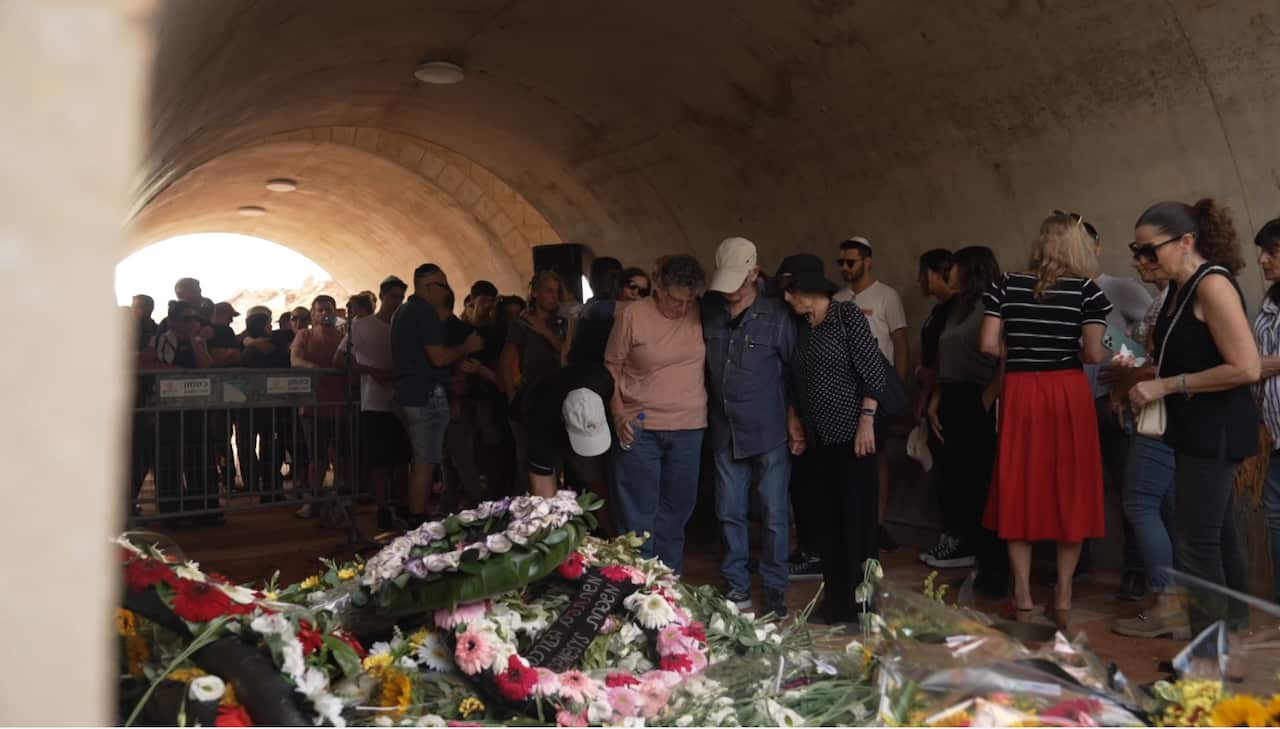 People huddled together at a funeral, in front of wreaths of flowers.