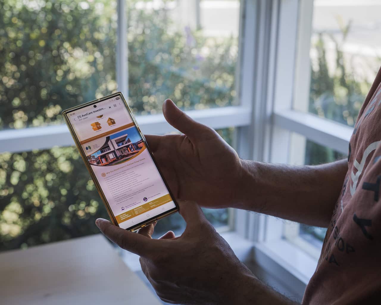 A person is holding a smartphone and scrolling through a website that displays information about real estate investment. The website features a photo of a modern house and text about "AusCare Investment," while a window with a view of green foliage is visible in the background.