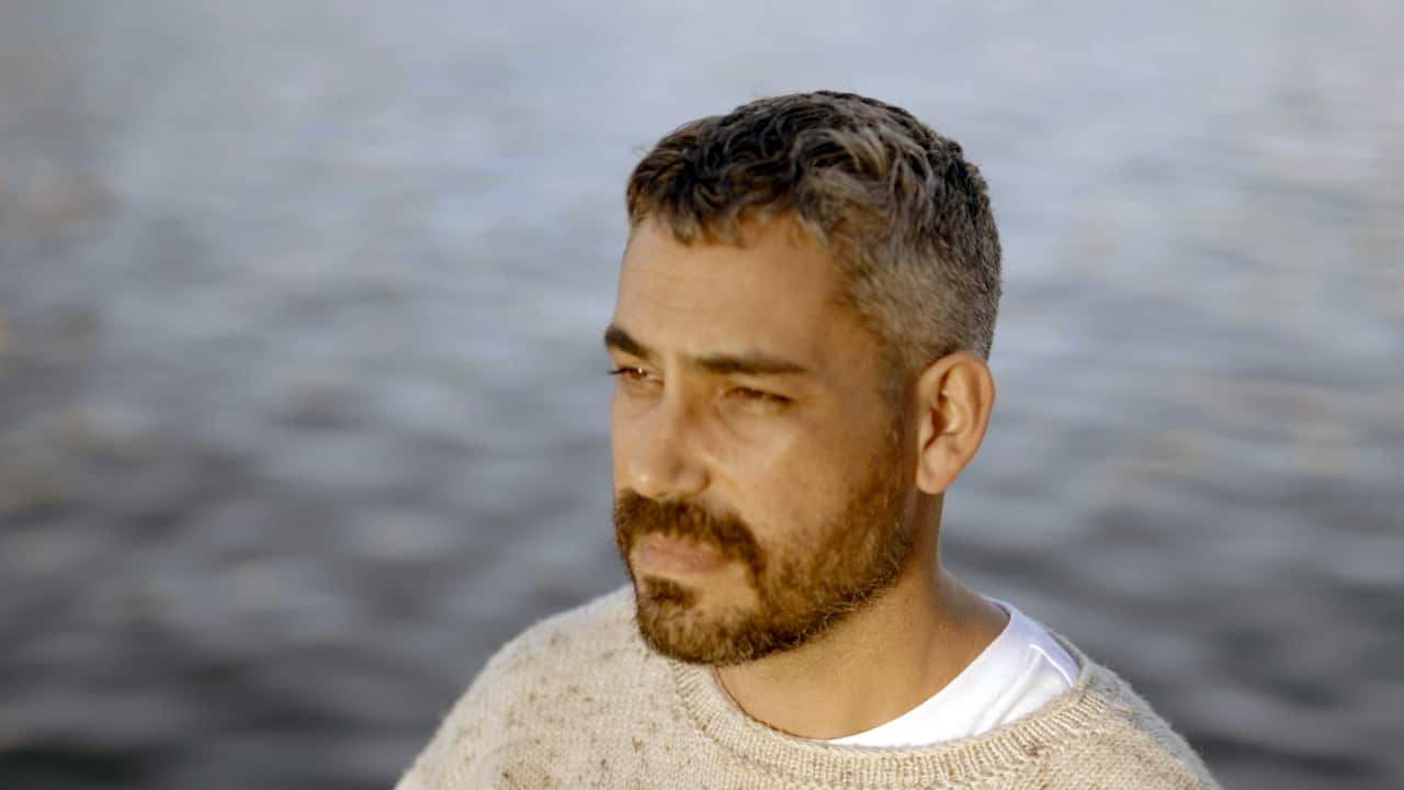 A man with a beard wearing a light-coloured jumper. There is water in the background