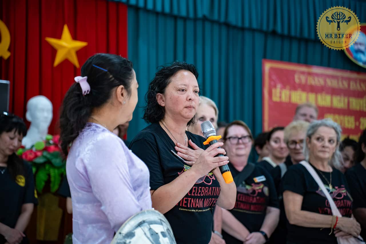 A middle-aged woman of Asian descent wearing a black t-shirt that reads 'Celebrating Vietnam Mothers' holds a microphone with her hand on her chest. Behind her, out of focus, is a stage with a curtain featuring the Vietnamese flag, and a group of people watching her.