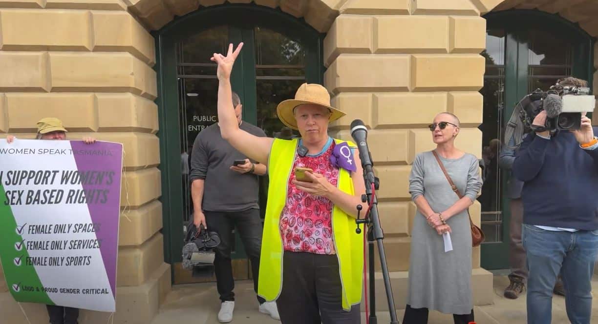 a woman in a high vis vest, standing in front of a sandstone building and wearing a large purple badge with the feminine symbol on it, holds her hand aloft with the peace symbol