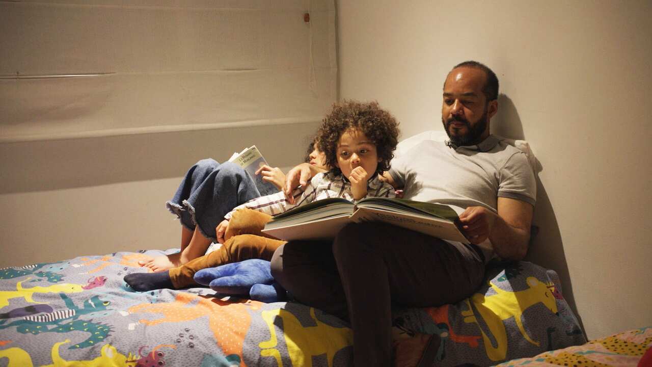 A man in a grey polo shirt sitting on a bed with two children and reading a book to a boy. A girl is lying next to them and reading a book