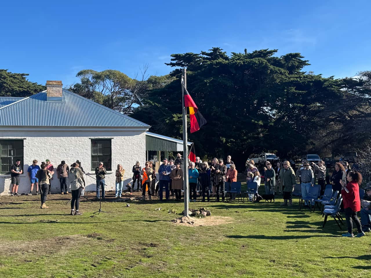 A group of people gathered around an Aboriginal flag on a flagpole outside
