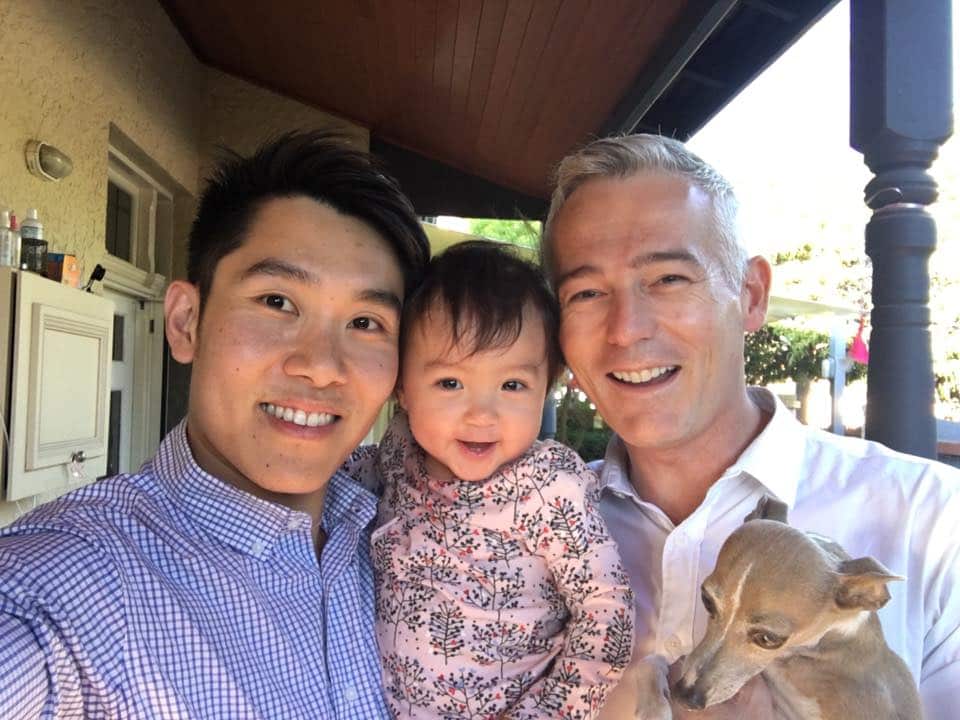 An Asian man and a Caucasian man taking a selfie with a toddler and a dog 