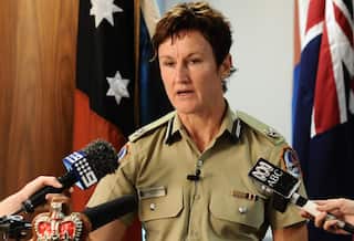 A woman in green police uniform speaking. There are media microphones near her.