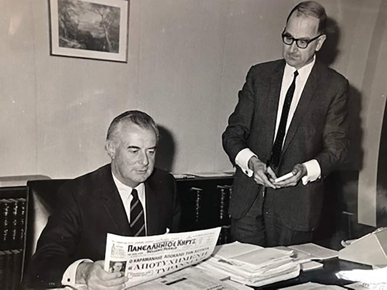 A black and white photograph shows Gough Whitlam seated at a desk reading a Greek newspaper while another man in a suit stands beside him.