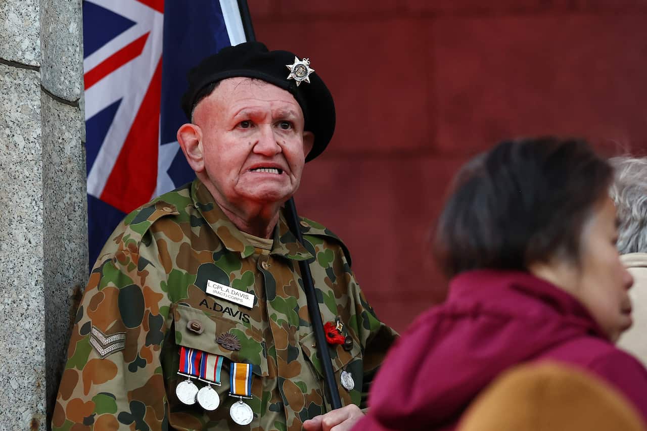 A man wearing camouflage uniform with medals on it and a black beret with a star badge on it. He is holding a large Australian flag on a pole.