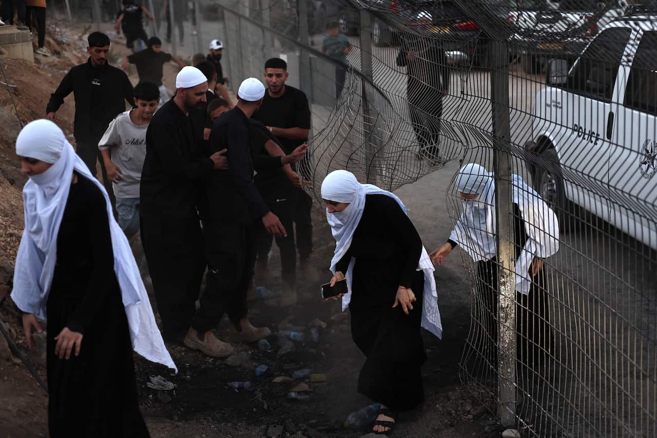 People, including women in traditional dress, pass through a bent fence while others gather nearby in a dusty area next to a police vehicle.