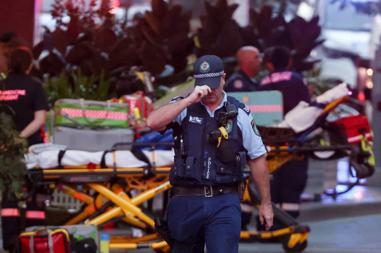 A NSW Police officer pulls the peak of his cap down over his eyes as he walks away from emergency services personnel and an ambulance stretcher