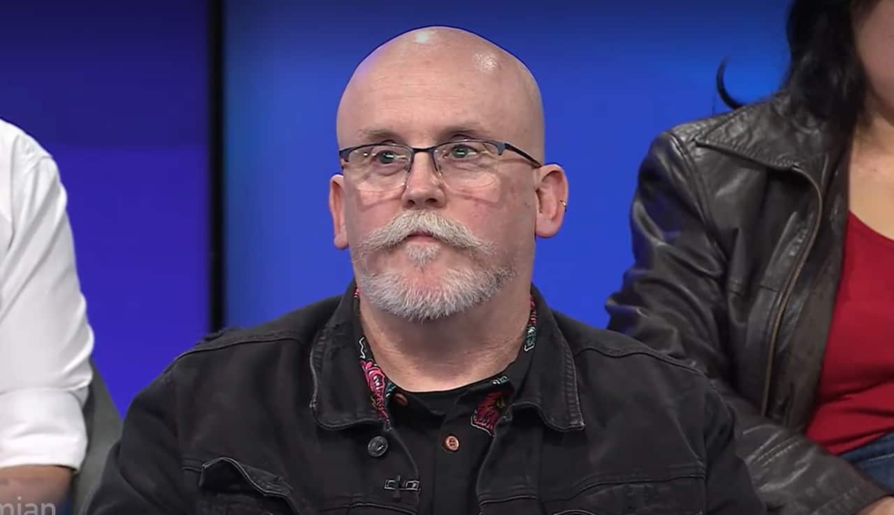 A middle-aged white man who is bald, wearing glasses, and a black jacket. He has a grey moustache and short beard around his chin, and an earring in his left face. He is sitting among a crowd of people. 