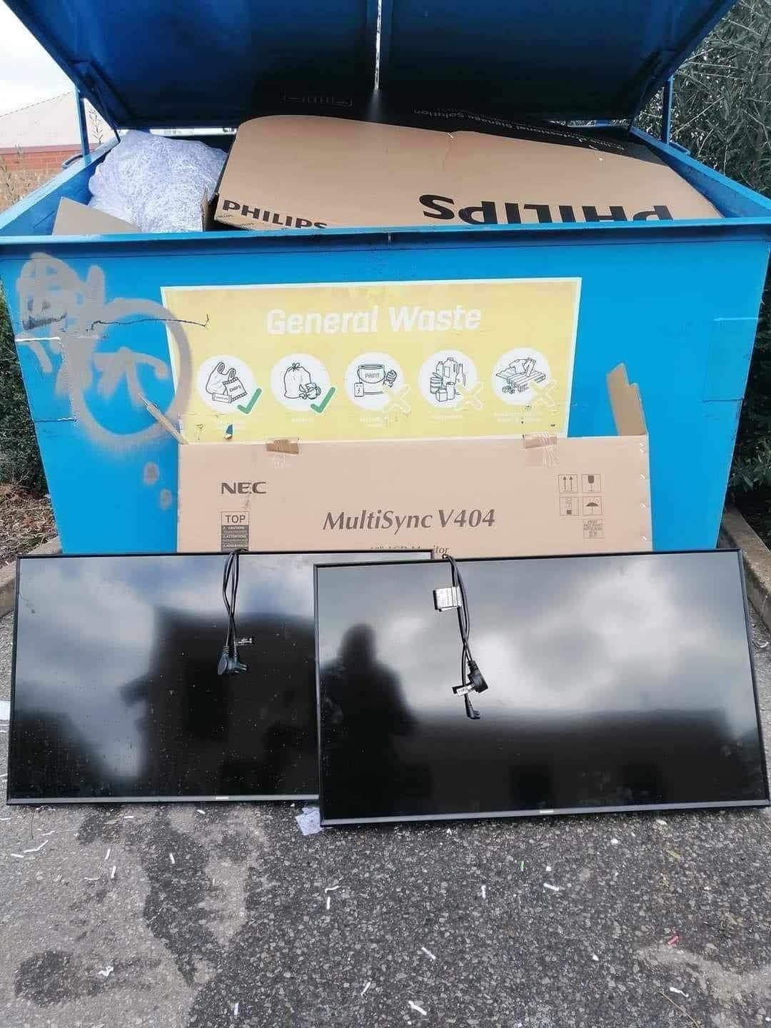 Two large TVs unplugged with a cable dangling outside a blue bin skip titled "general waste"