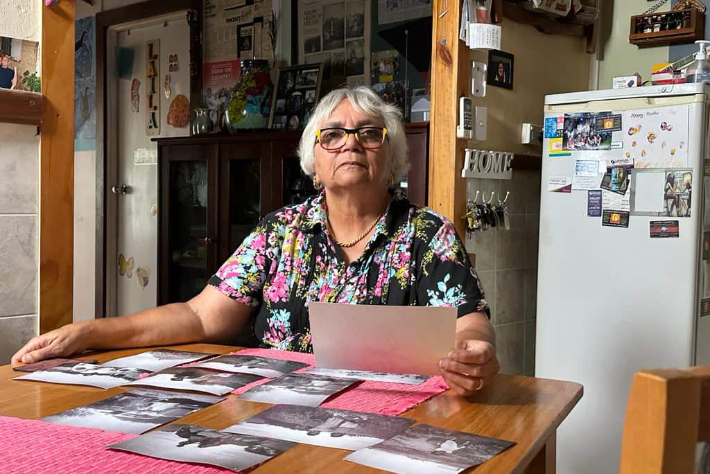 A woman in glasses sits at a kitchen table. She holds a photograph in her hand. More black and white photographs are spread out on the table. 