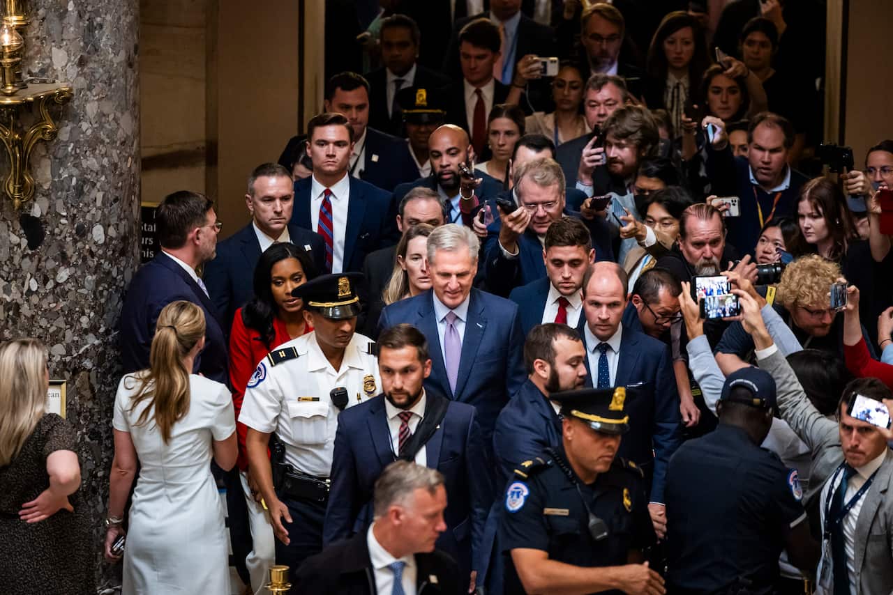 A man wearing a suit and tie surrounded by police and journalists as he walks.