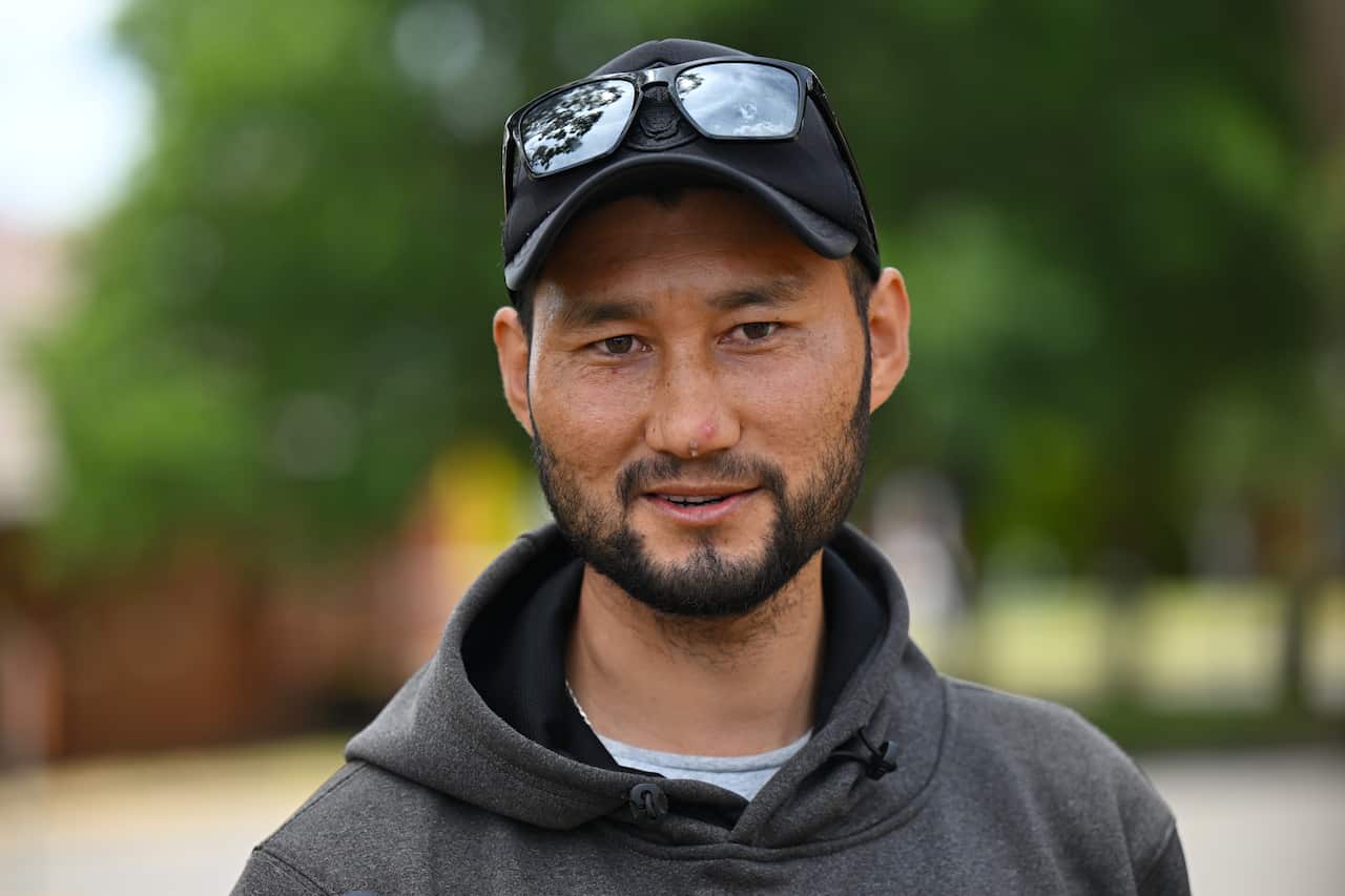 A man wearing a dark grey hoodie, black cap, and sunglasses atop the cap is smiling.