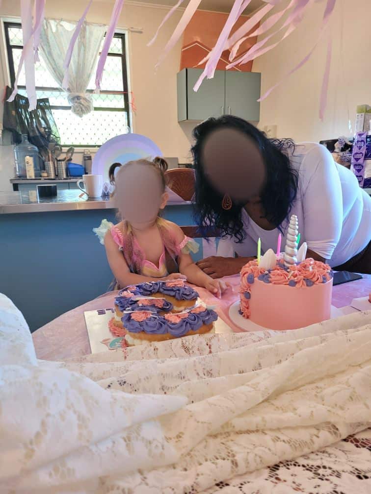 An Indigenous woman with her granddaughter, seated at a kitchen table with a birthday cake. Their faces have been blurred.