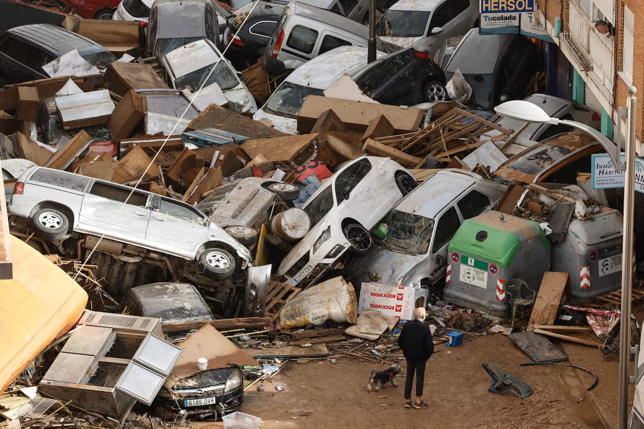 An elderly man stands in front of a pile of cars and debris