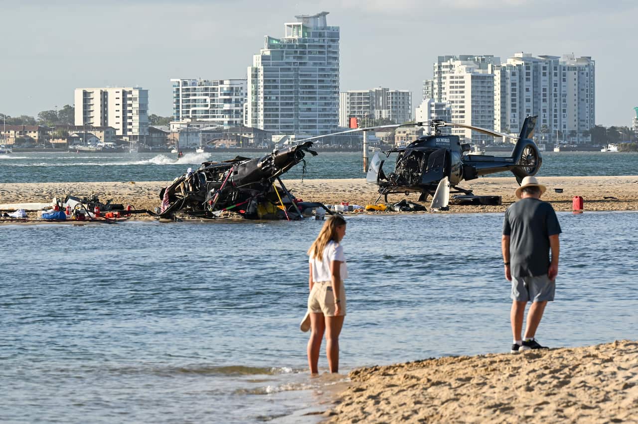 Two people walk along the beach in the foreground, beyond them on a sand bank are the broken wreckages of two helicopters.