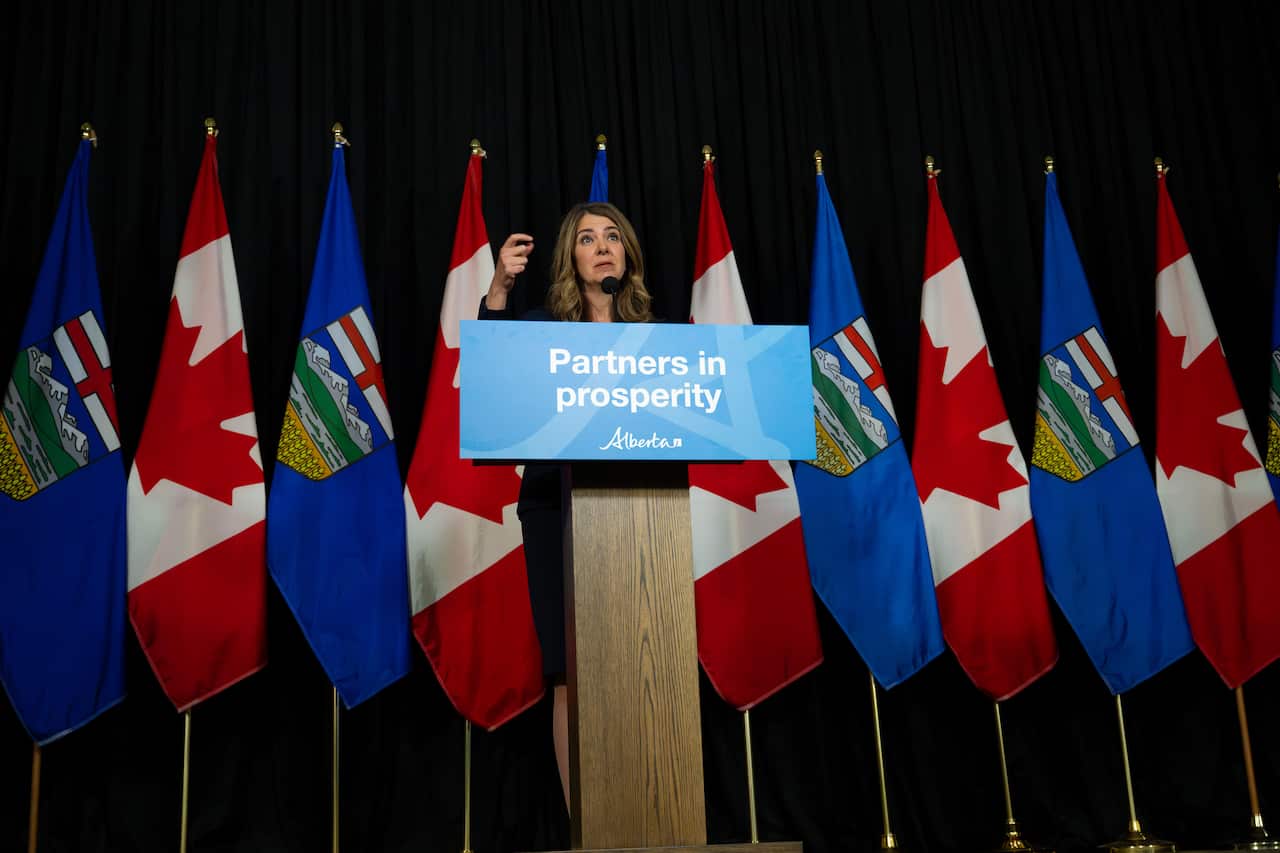 Danielle Smith stands at a podium in front of a row of Canadian and Albertan flags.