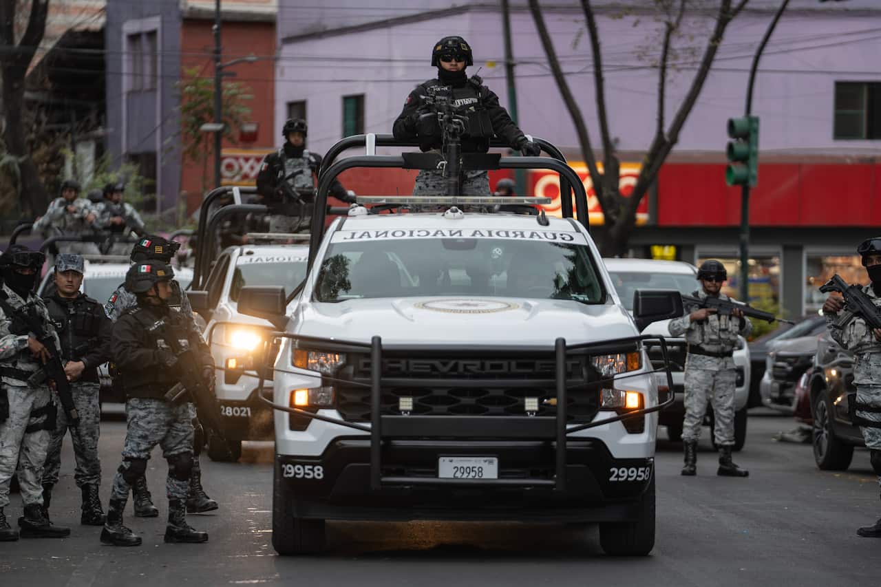 Armed National Guard officers in tactical gear stand around and atop white patrol trucks with mounted weapons on a city street.
