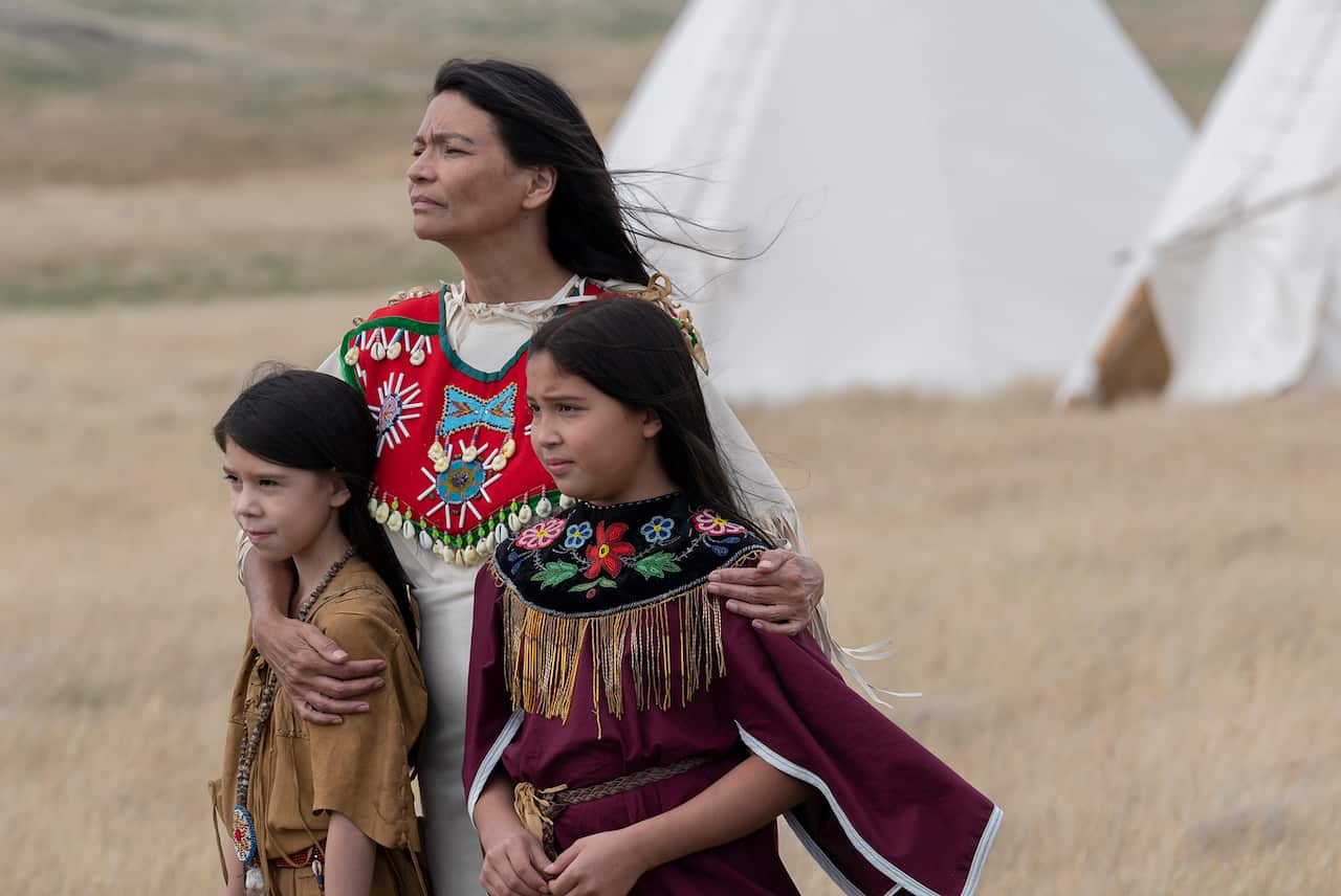 An Indigenous woman stands on a grass plain, with her arms around two young girls. A white tent can be seen in the background. 