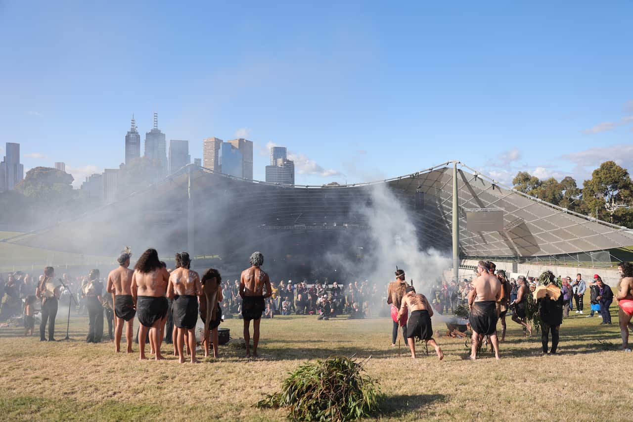 men in traditional paint and dress stand outside on the grass in front of an amphitheatre, with the melbourne city skyline in the background. 