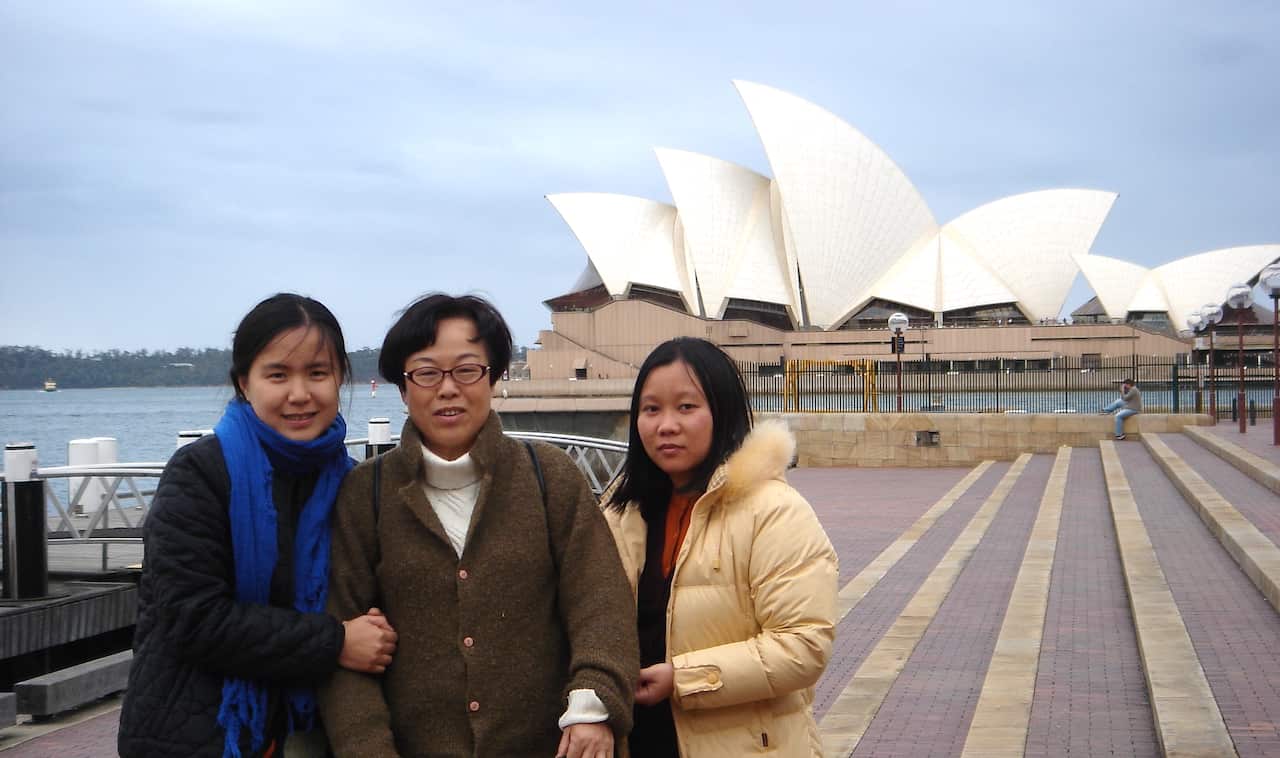 An elderly woman, flanked by two young girls on either side, is pictured in front of the Sydney Opera House.