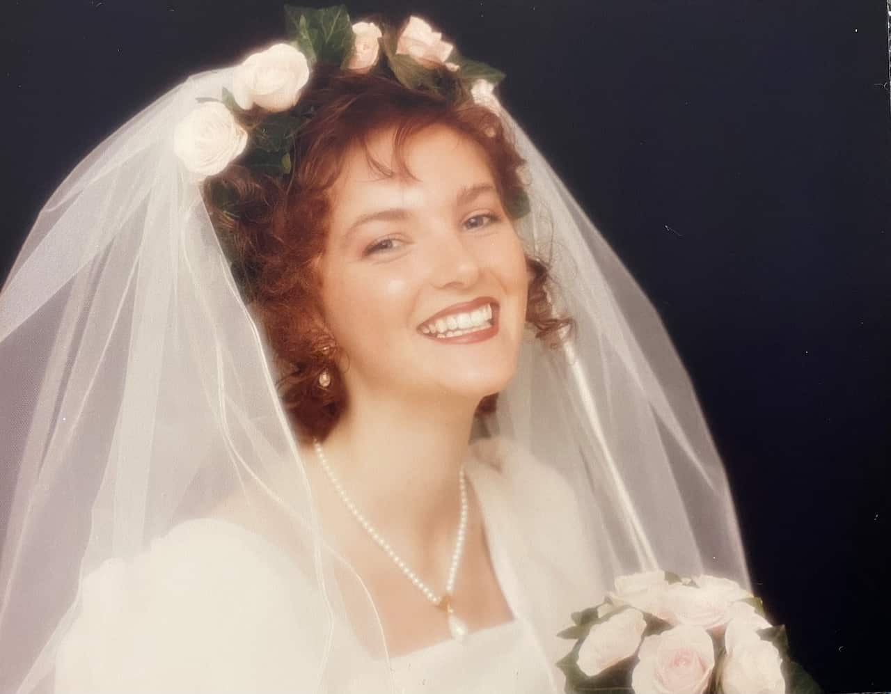 A head-and-shoulders shot of a smiling bride in a white dress. She is wearing a white pulled-back veil with and holding a small bunch of pink and white roses. There are pink and white roses in a band on her head. 