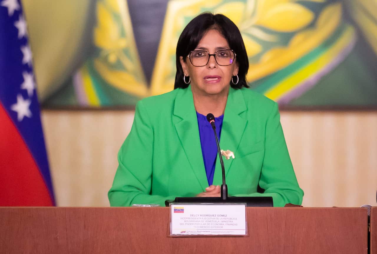 A woman in a green blazer, standing at a podium, is speaking.