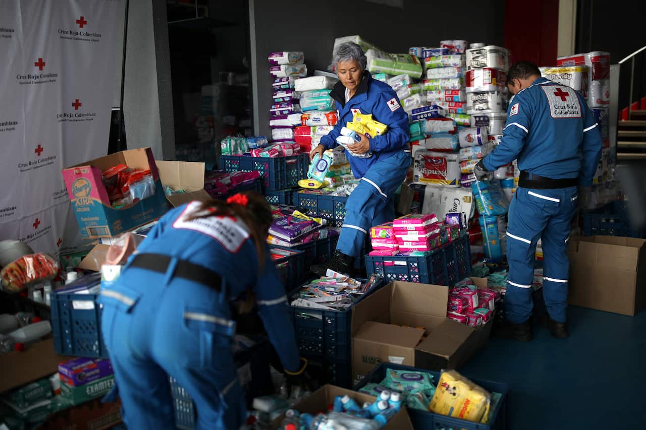 Three Red Cross workers wearing blue jumpsuits gather food and water supplies for earthquake survivors in a tent.