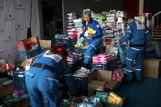 Three Red Cross workers wearing blue jumpsuits gather food and water supplies for earthquake survivors in a tent.