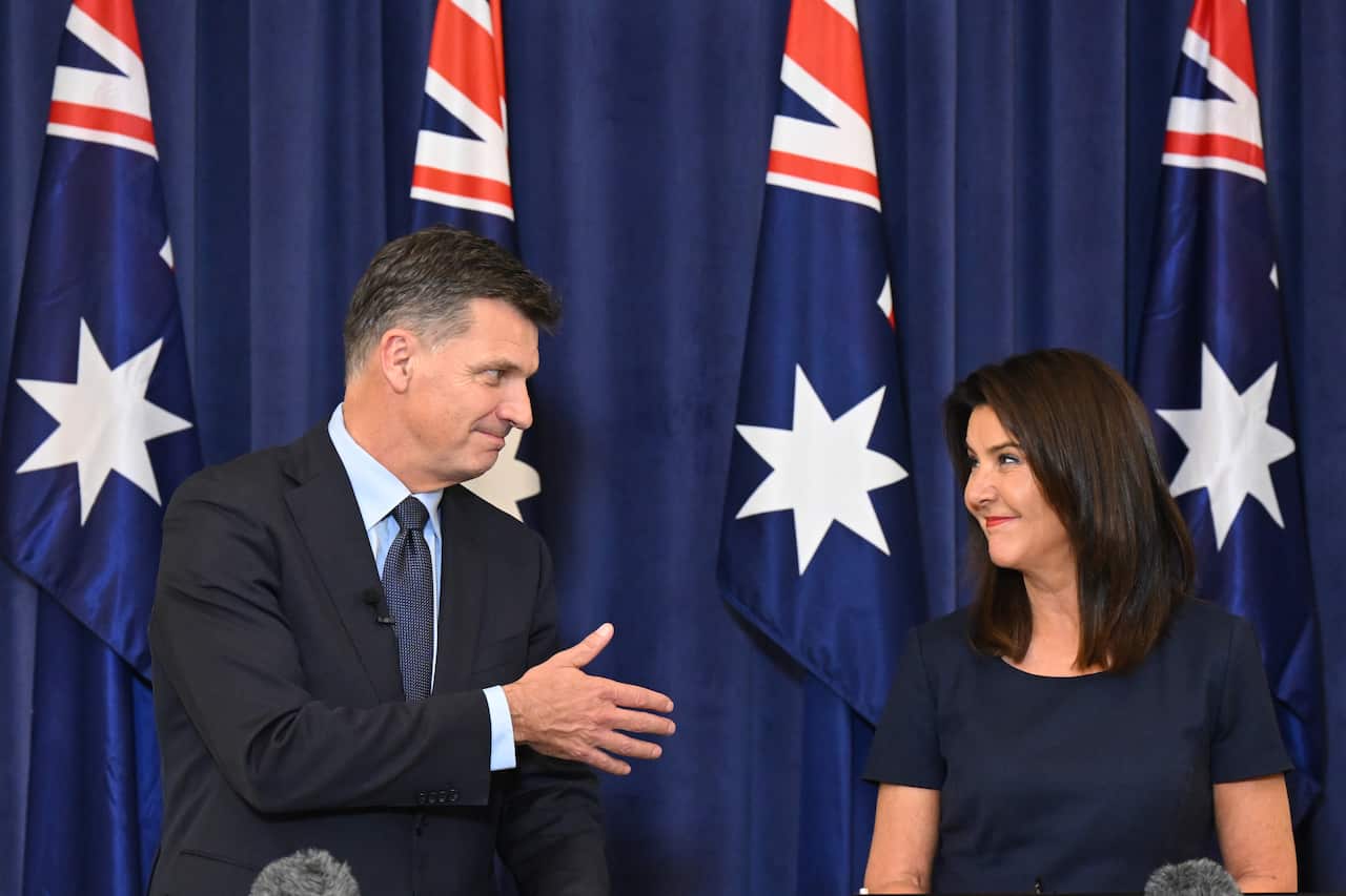 Angus Taylor, wearing a suit, extends his hand towards Jane Hume, who is wearing a dark top. There are Australian flags in the background