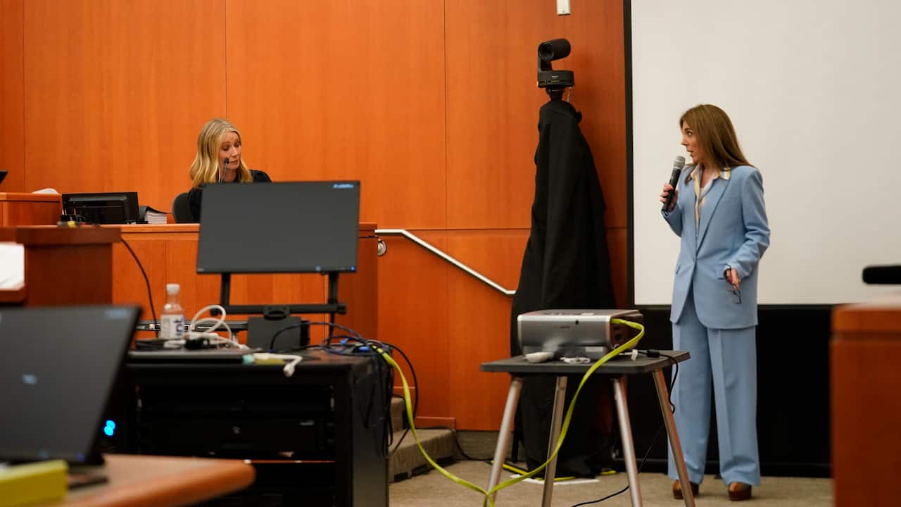 A woman seated in the witness stand in a courtroom listens while another woman, who is standing, speaks to her.