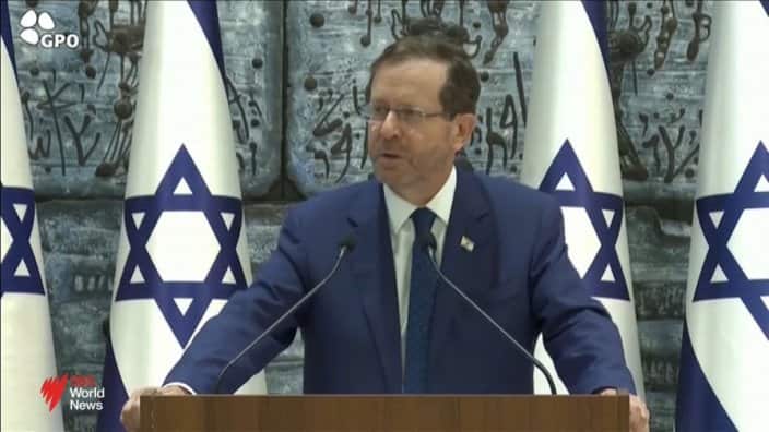 A man in a suit and tie speaking at a lectern. There are Israeli flags behind him