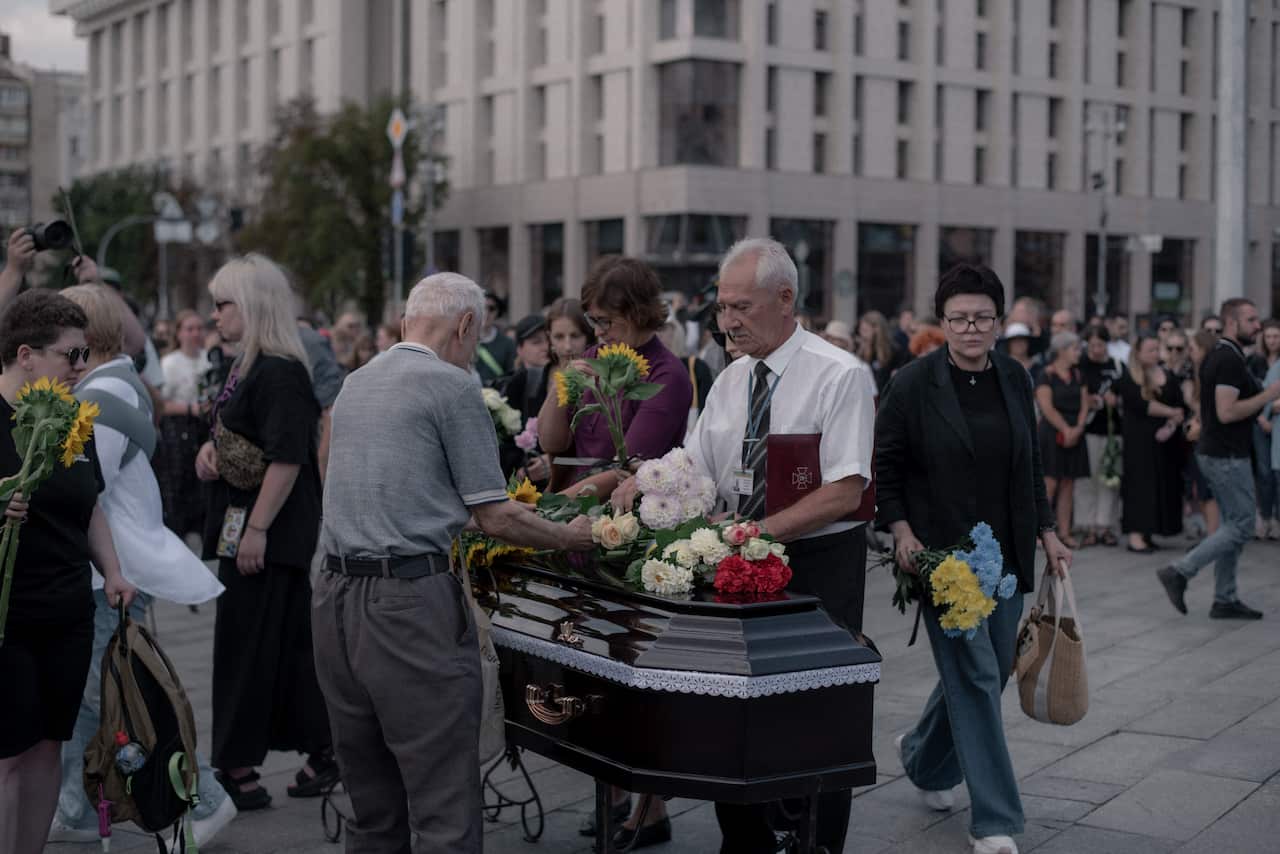 People stand outdoors while two elderly men place flowers on a closed coffin